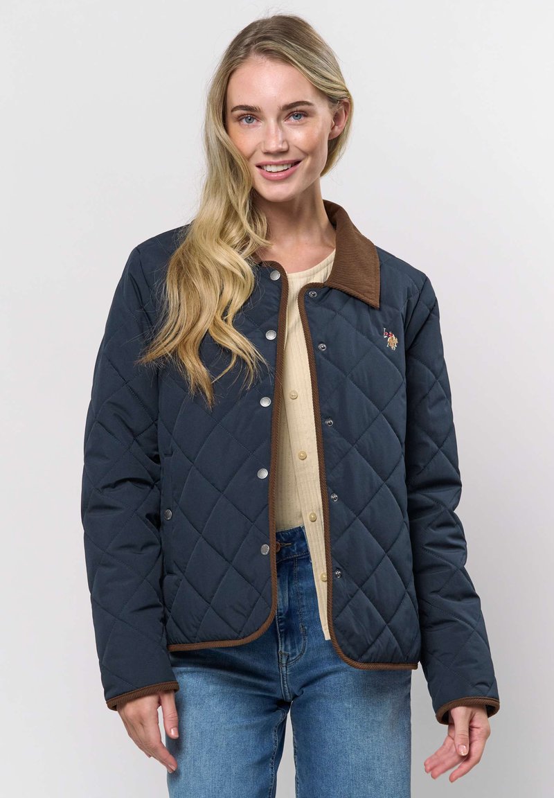 Jonge vrouw met lang blond haar, gekleed in een marineblauwe doorgestikte jas met bruine bies, een beige blouse met knopen en een blauwe spijkerbroek, glimlachend naar de camera.