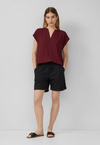 Bordeaux textiel top met V-hals en korte mouwen, gecombineerd met zwarte shorts met opgerolde zoom en zwarte platte sandalen. Eenvoudige casual outfit.