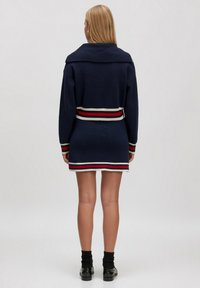 Navyblaues Strickkleid mit Kragen, gestreiftem Taillenbund in Rot- und Weißtönen sowie langen Ärmeln. Glatte Textur, figurbetonte Silhouette.