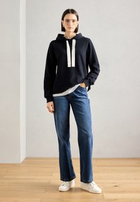 Juoda sportinė palaidinė su kapisonu ir kreminėmis virvutėmis, derinama su mėlynomis plačiomis kelnėmis ir baltomis sportbačiais. Minimalistinis, kasdieniškas dizainas.