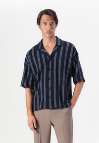 Camisa de botones de manga corta en azul marino oscuro con rayas verticales en azul claro, corte relajado y cuello plano. Textura de tela suave.