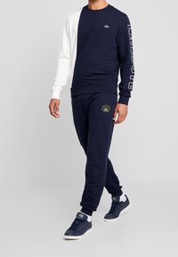 Felpa blu navy con manica bianca, caratterizzata da scritte audaci, abbinata a pantaloni jogger blu navy coordinati e sneaker blu navy con dettagli bianchi.