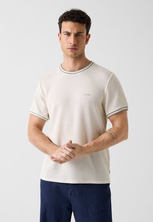 Uomo con capelli corti e ricci, indossa una maglietta beige a maniche corte con bordi grigi e pantaloni blu scuro, con le mani giunte davanti.