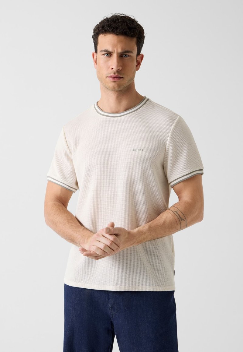 Homme aux cheveux courts et bouclés portant une chemise en maille beige à manches courtes avec un bord gris et un pantalon bleu foncé, les mains jointes devant lui.