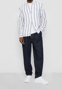 Camicia in lino a righe bianche e blu, con taglio rilassato e chiusura frontale con bottoni; abbinata a pantaloni in denim scuro e sneakers bianche.