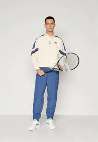 Meeste spordivorm, mis sisaldab pruunist kreemikast jakki siniste ja punaste aktsentidega, mis on kombineeritud siniste jooksupükstega ja valgete tossudega, hoides tennisereketit.