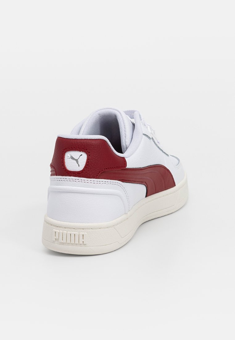 Sneakers en cuir blanc avec des accents rouges, talon rembourré et semelle en caoutchouc texturé. Présente un logo Puma bien visible au niveau du talon et sur le côté latéral.