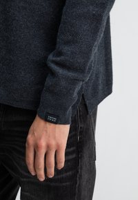 Dunkelgrauer Strickpullover mit weicher Textur, seitlichem Schlitz und einem Markenlabel am Bündchen. Modell trägt dunkle Jeans.