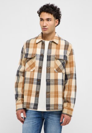Young man wearing a brown, beige, and gray plaid button-up shirt over a white t-shirt and blue jeans, looking slightly to the side.