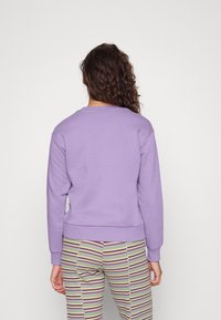 Persona con capelli ricci di spalle, che indossa un top a maniche lunghe lilla e pantaloni a strisce colorate, su uno sfondo chiaro e uniforme.