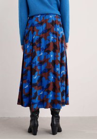 Donna che indossa una gonna marrone con grandi stampe floreali blu, stivali neri con tacco e un maglione blu, in piedi su un pavimento grigio.