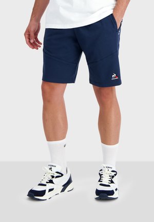 Pantalones cortos deportivos de color azul marino con una textura suave, presentan un logo bordado y bolsillos laterales. Combinados con calcetines blancos hasta el tobillo y zapatillas.
