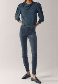 Kvinna iklädd blå puffärmad blus, hög midje slim-fit blå byxor och svarta loafers, stående med händerna i bakfickorna.