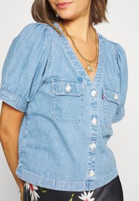Chemise en denim bleu clair avec manches courtes bouffantes, col en V, fermeture par boutons et deux poches poitrine. Présente des coutures contrastées et un ourlet arrondi.