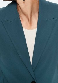 Tyrkysový blejzr s výrazným límcem a jedním černým knoflíkem, nošený přes lehký texturovaný top, s čistým a upraveným designem.