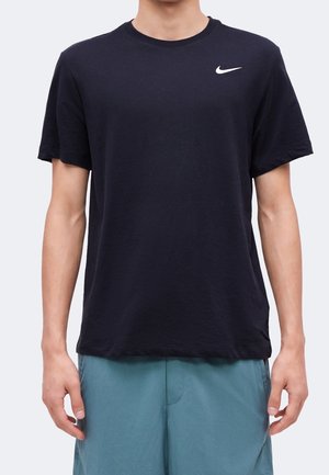 Persona che indossa una semplice t-shirt Nike nera con logo bianco sul petto e pantaloncini turchesi, in piedi su uno sfondo chiaro.