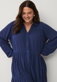 Vestido azul marino con botones en la parte delantera, cuello con solapa, mangas largas y diseño en capas. Hecho de tela ligera con una sutil textura.