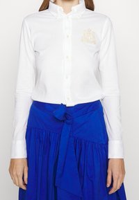 Vit långärmad skjorta med knappar och krage, med gyllene broderi på vänster bröst, tillsammans med en blå kjol med knyt i midjan.