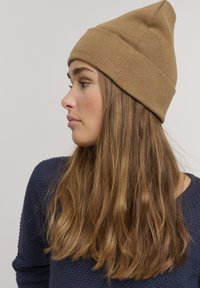 Gorro de punto beige con una parte superior puntiaguda. El modelo tiene el cabello castaño largo y ondulado y lleva un suéter texturizado azul marino. Diseño sencillo y coherente.
