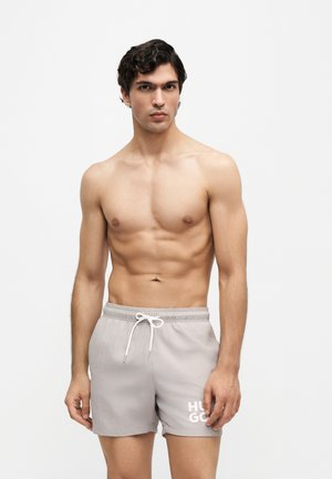 Joven en forma con cabello rizado oscuro, vistiendo pantalones cortos grises claros con cordón, de pie frente a un fondo blanco liso.