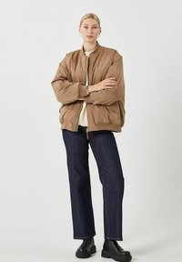 Chaqueta bomber marrón con cremallera frontal, corte oversize, cuello acanalado, llevada sobre un suéter crema y pantalones de mezclilla oscuros, combinada con botas negras.