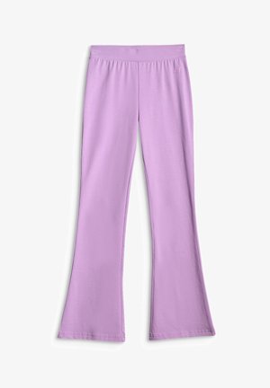 Une jeune fille souriante porte un sweat-shirt rose clair, un pantalon évasé lavande et des baskets blanches sur un fond gris uni.