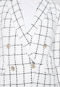 White double-breasted blazer in textured fabric with black grid pattern, featuring gold-tone buttons and sharp lapels.