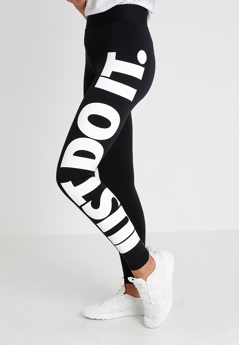 Nogi ubrane w czarne legginsy z napisem "JUST DO IT." dużymi białymi literami, w połączeniu z białym obuwiem sportowym na jednolitym tle.