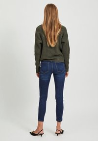 Pull en maille vert foncé, jean skinny bleu avec poches arrière, et mules noires à talons bas. Cheveux longs tombent sur les épaules.