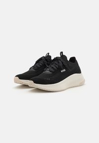 Černé sportovní tenisky s texturovaným pleteným svrškem, kontrastními černými tkaničkami a silnou, drážkovanou béžovou podrážkou. Logo značky viditelné na boku.