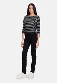 Czarno-biała, w paski bluzka z rękawem 3/4, połączona z czarnymi, dopasowanymi dżinsami i białymi trampkami, stojąca na jednolitym tle.