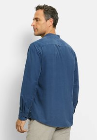 Blaues Langarmhemd mit subtiler Textur, Stehkragen und Knopfmanschetten. Die Rückseite hat einen zentralen Einsatz und einen abgerundeten Saum.