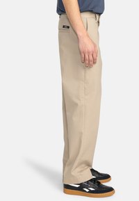 Pantalons en coton beige avec une coupe droite, dotés d'une poche arrière et portés avec des baskets noires. Texture lisse et coupe classique.