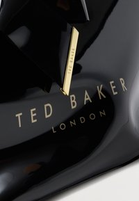 Γυαλιστερό μαύρο παπούτσι με χρυσό λογότυπο Ted Baker London και αντίστοιχο χρυσό ορθογώνιο διακοσμητικό στο πάνω μέρος.
