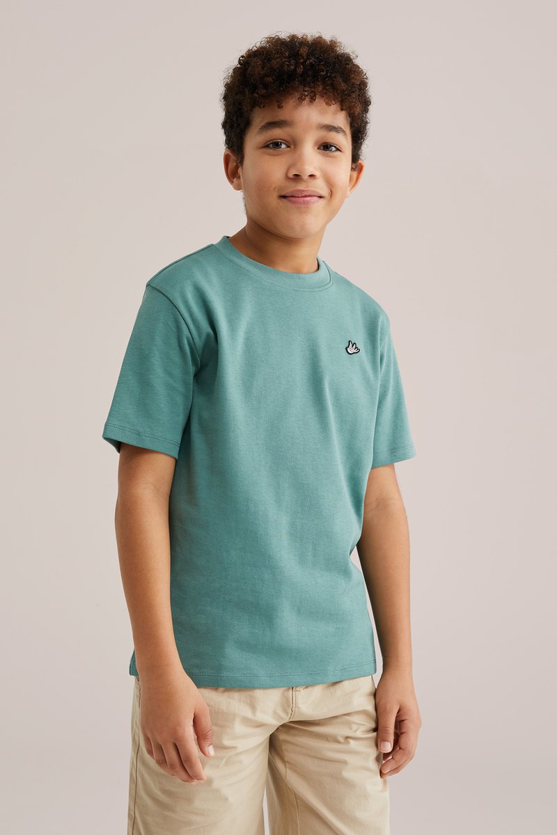 Zēns ar cirtainiem matiem, valkājot zaļgani zilu īsroku krekliņu un bēšas šortus, stāvot pret vienkāršu neitrālu fonu, nedaudz smaidot.