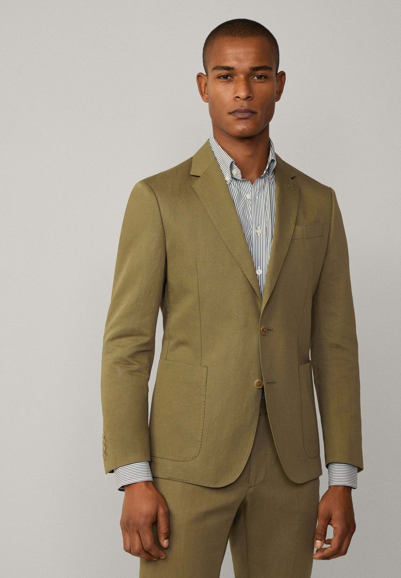 Olivgrüner Blazer mit zwei Knöpfen, zwei vorderen Taschen und Reverskragen. Getragen über einem gestreiften Hemd mit Kragen- und Manschettendetails.