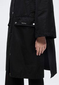 Χέρι που φοράει μαύρο παλτό με μακριά μανίκια, με το λογότυπο Barbour στο κούμπωμα της τσέπης, πάνω σε απλό λευκό φόντο.