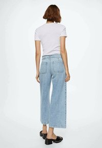 Jeans de pierna ancha en azul claro con cintura alta, que presentan dos bolsillos traseros y un dobladillo sin rematar. Combinados con una camiseta blanca de manga corta lisa y sandalias de tacón negras.