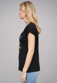 Svart bomulls-T-shirt med rund hals och korta ärmar, med ett grafiskt tryck, tillsammans med ljusblå jeans. Sido- eller profilvy.