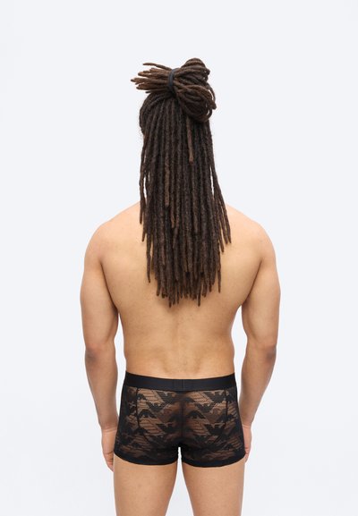 Άνδρας με μακριές κοτσίδες δεμένες, φοράει μαύρα boxer σλιπ με σχέδια από δίχτυ, στέκεται με την πλάτη προς την κάμερα μπροστά σε ένα απλό φόντο.