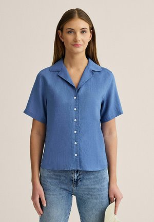 Kvinde iført kortærmet blå skjorte med knapper og jeans, holder en hvid clutch og står foran en ensfarvet beige baggrund.