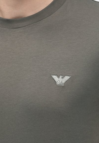 Šedé bavlnené tričko s okrúhlym výstrihom a malým, vyšívaným logom orla v striebornej farbe na ľavej strane hrudníka. Hladká textúra.