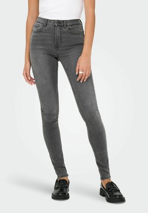 Jean skinny gris taille haute avec une texture lisse et un design à cinq poches. Associée à des mocassins noirs, le modèle pose devant un fond simple.