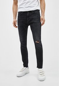 Jeans negros de corte ajustado hechos de denim, con un detalle de rodilla rasgada y un acabado desgastado, combinados con zapatillas blancas.