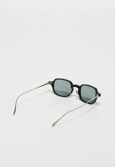 Schwarze quadratische Sonnenbrille mit grünen Gläsern und dünnen goldenen Metallbügeln. Die Details umfassen schlanke Bügelspitzen und ein glänzendes Finish am Rahmen.