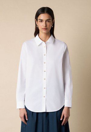 Vrouw met steil donker haar draagt een wit overhemd met lange mouwen en een donkerblauwe geplooide rok, staand tegen een beige achtergrond.