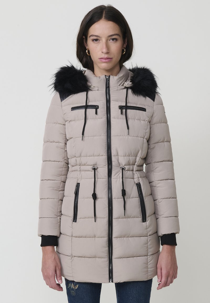Polstret beige vinterjakke med en sort faux fur hætte, lodrette lynlåslommer og en snoretræk i taljen. Har sorte accentdetaljer.
