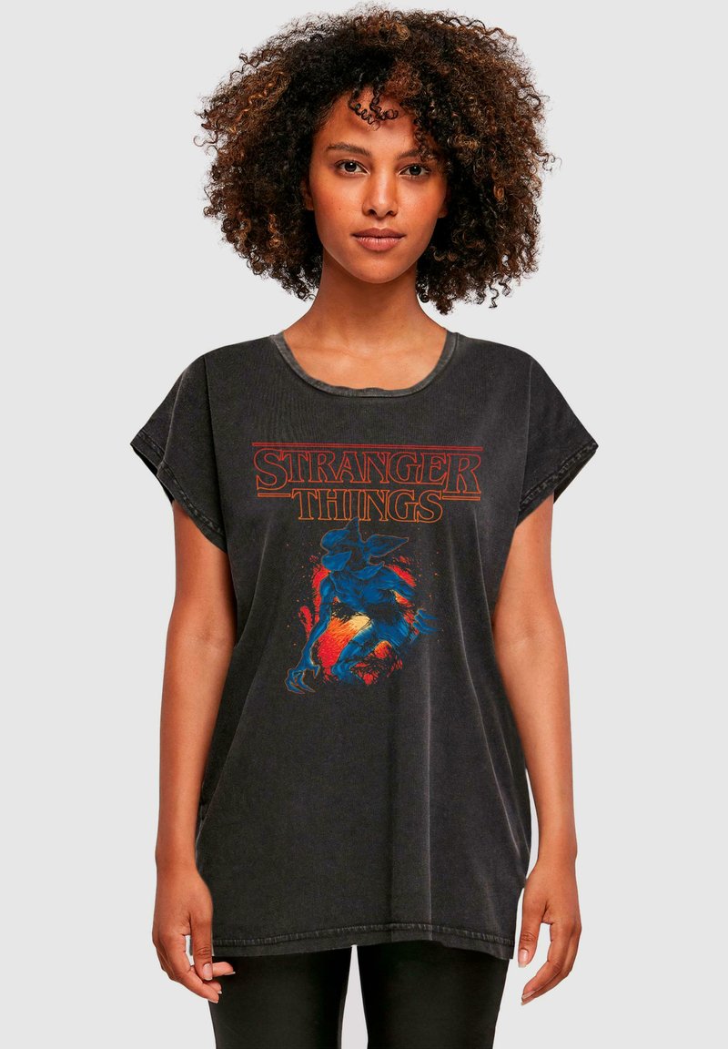 Juodas per didelis marškinėliai su "Stranger Things" logotipu raudonai ir iliustracija mėlyno padaro su ryškiu fono detalėmis.
