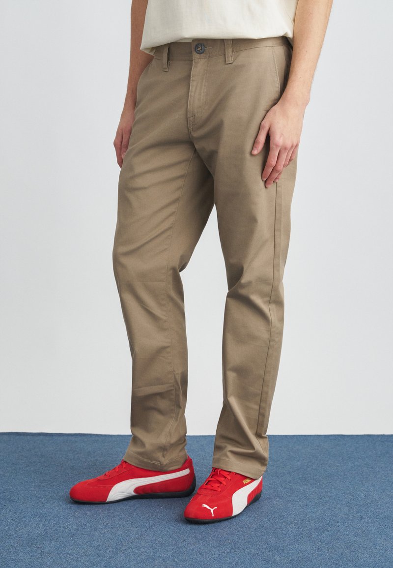 Μπεζ βαμβακερό παντελόνι με ευθεία γραμμή, διαθέτει κλείσιμο με κουμπί και τσέπες, συνδυασμένο με κόκκινα αθλητικά παπούτσια.