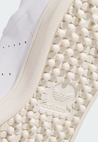 Vit sneaker med en texturerad läderovandel, perforeringsdetaljer och en gummisula med upphöjda cirkulära mönster och logotyp.